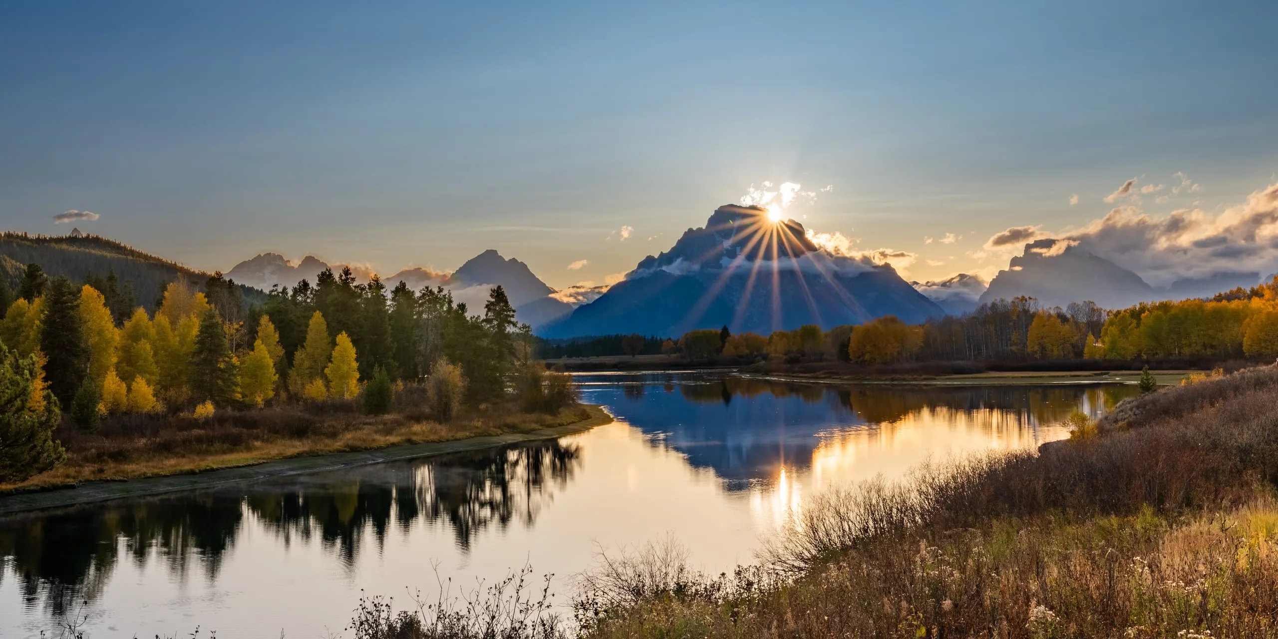 The sun breaking over Mount Moran at Oxbow Bend, with fall foliage and a perfect reflection in the still Snake River