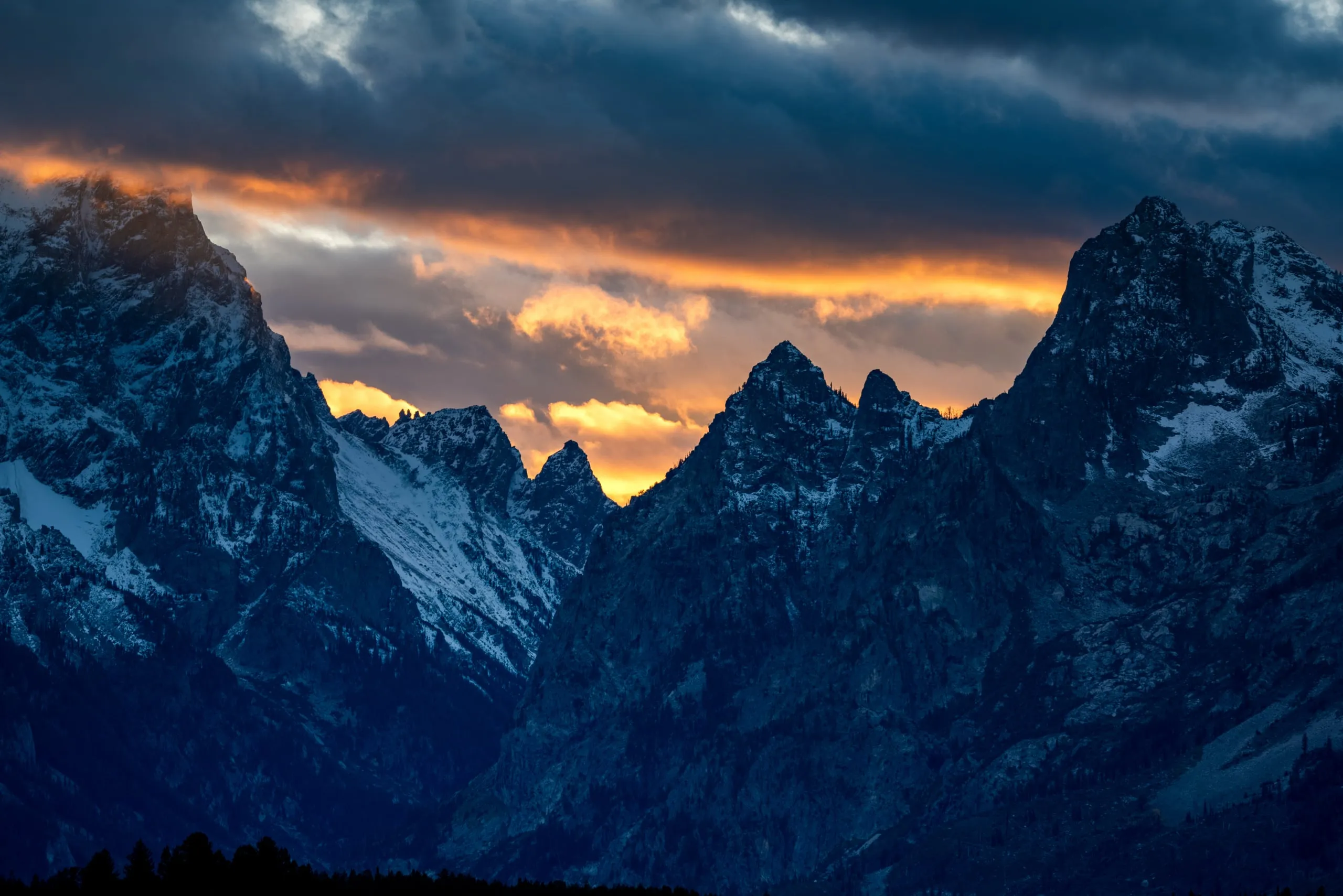 Dramatic orange sunset light breaking through storm clouds above the jagged, snow-dusted Teton peaks
