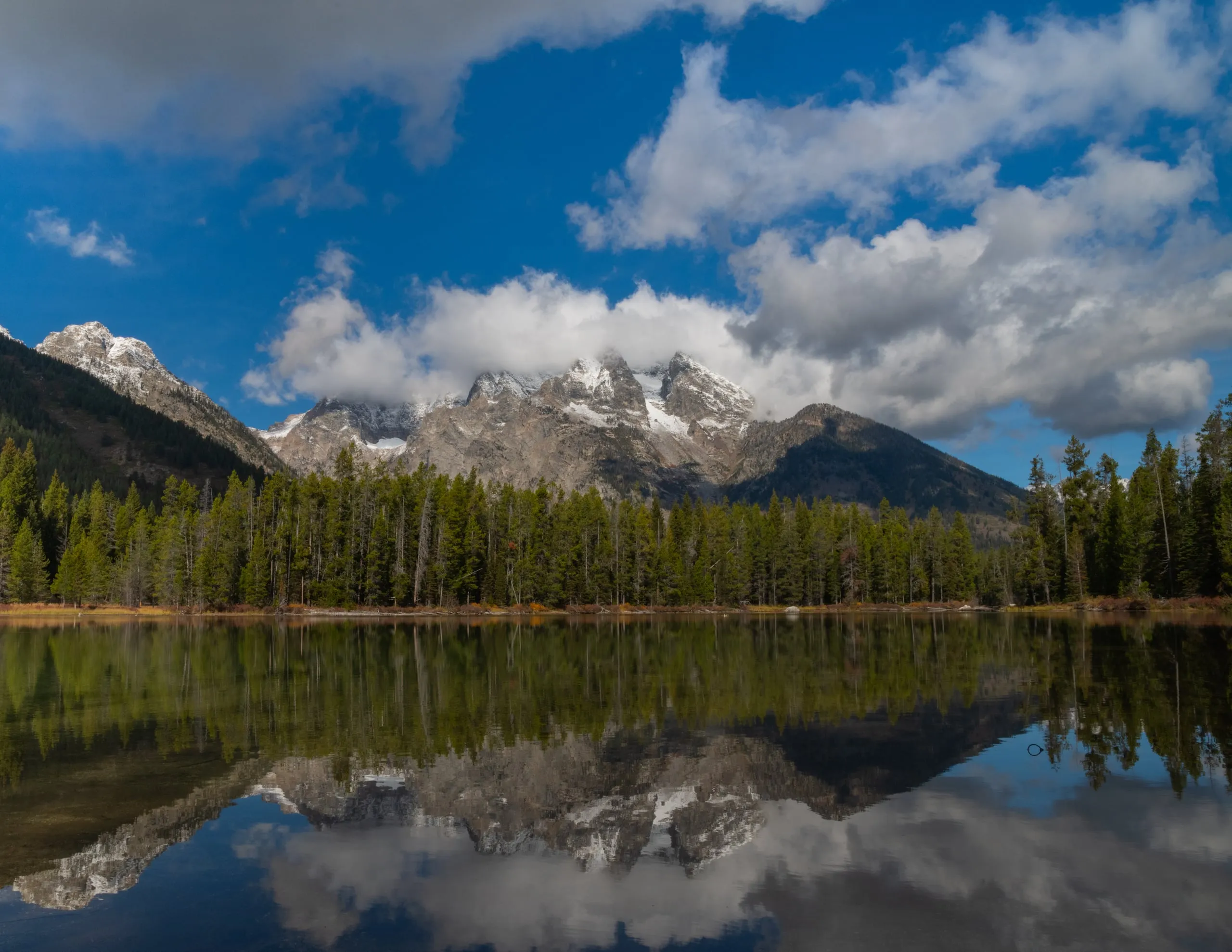 Snow-dusted Teton peaks reflected in a still lake, with clouds parting overhead to reveal the summits