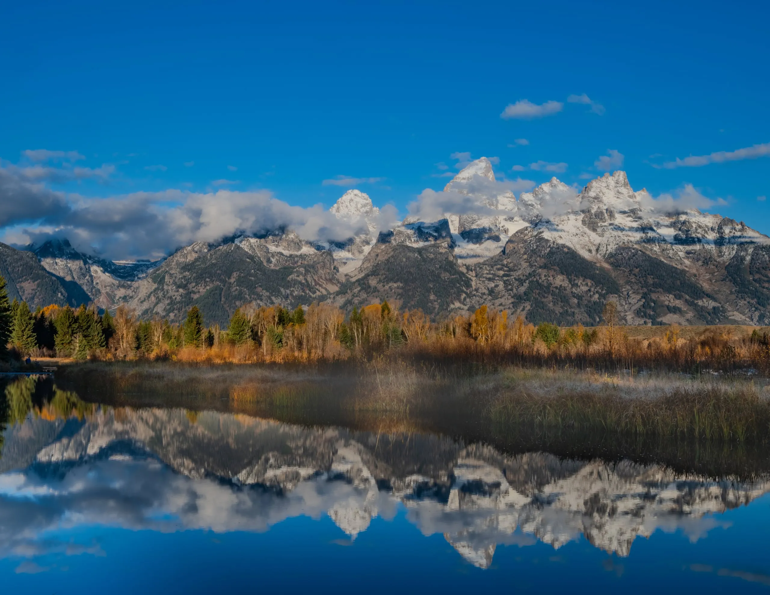 The Teton range reflected in a still beaver pond at dawn, mist rising off the water through golden fall foliage