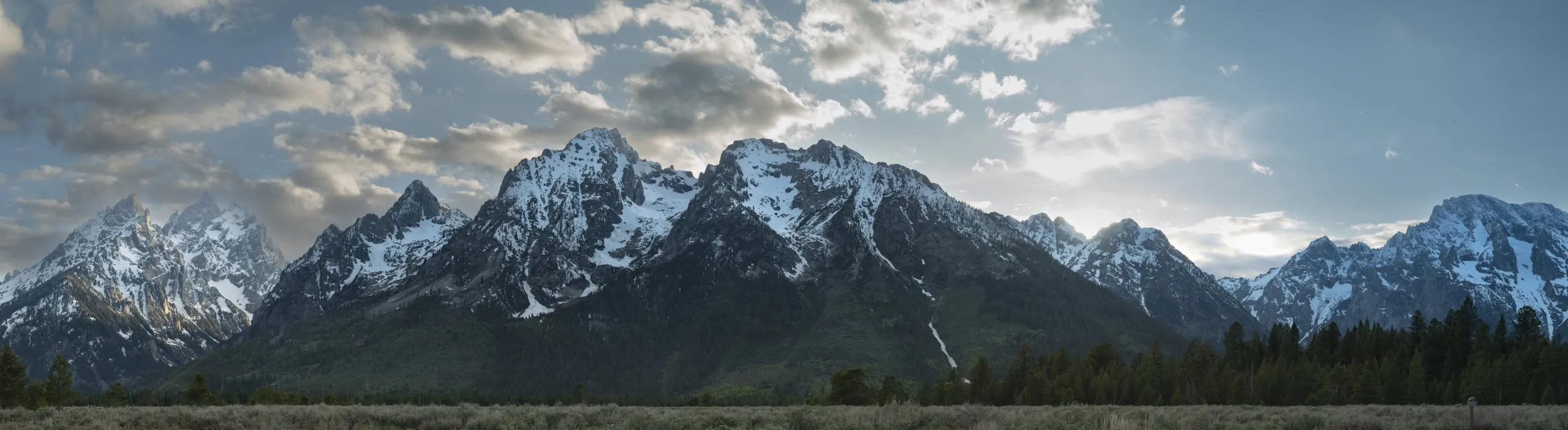 A wide panorama of the entire snow-capped Teton range under a partly cloudy sky, sagebrush flats in the foreground