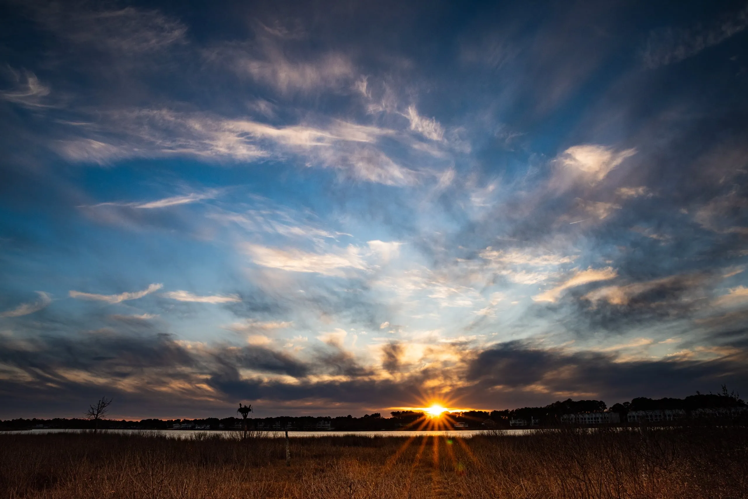 The sun setting in a starburst over a bay marsh, wisps of cloud lit gold across a wide sky, an osprey nest platform silhouetted in the foreground